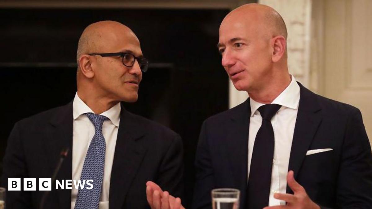 Microsoft CEO Satya Nadella (left) and Amazon founder Jeff Bezos (right), both wearing black suits over white shirts and sporting neck ties, having a conversation at an event in the US