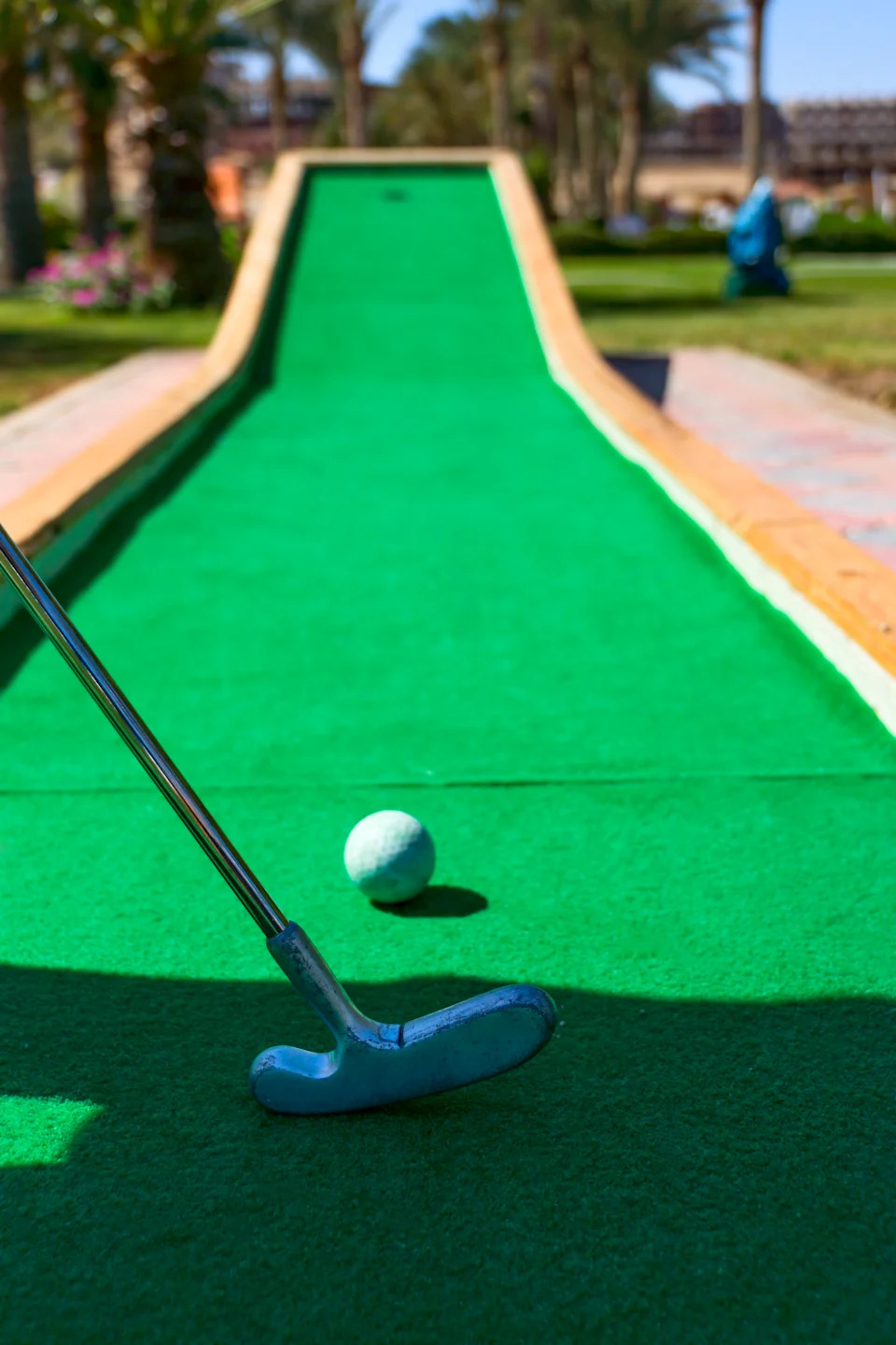 Golf ball on a miniature golf course, near a putter ready to strike, in a sunny outdoor setting