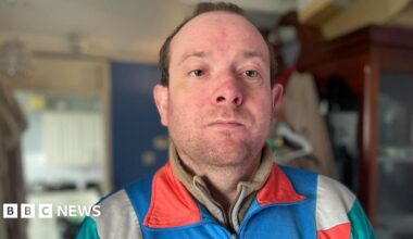 A man with dark thinning hair and a beard wears a blue tracksuit top and underneath a beige fleece