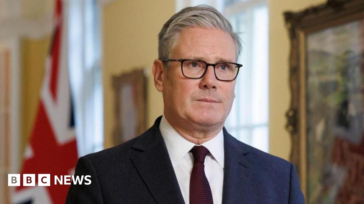 A young man with dark curly brown hair standing by the side of a road. He is wearing a black t-shirt and behind him are yellow crops and blue skies.