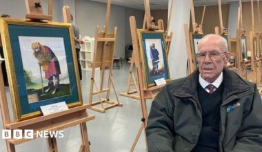 Dick Scott, with balding grey hair and a moustache, wearing glasses, a green coat, navy jumper, white shirt and tie, sits in a chair next to a series of watercolour paintings on a wooden easels.