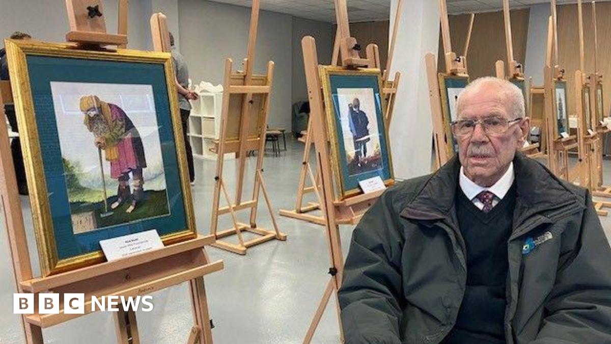 Dick Scott, with balding grey hair and a moustache, wearing glasses, a green coat, navy jumper, white shirt and tie, sits in a chair next to a series of watercolour paintings on a wooden easels.