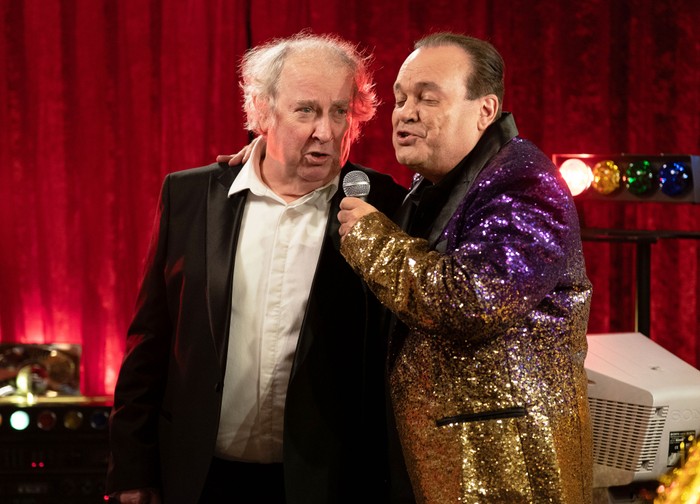 Nigel, in a black suit and white shirt, sings karaoke with Barry, who is in a sequinned gold and purple blazer in EastEnders
