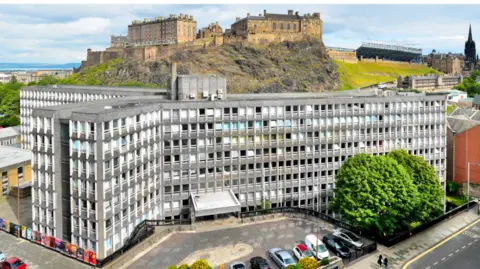 Hendersonherd An aerial view of Argyle House, which is grey with lots of windows, designed in the brutalist style. Behind it is Edinburgh Castle.