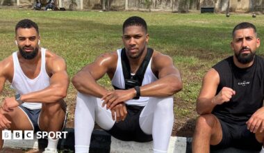 Anthony Joshua sits between Sina Ghami and Latif Ayodele as all wear training kit, with a grassy area behind the three men