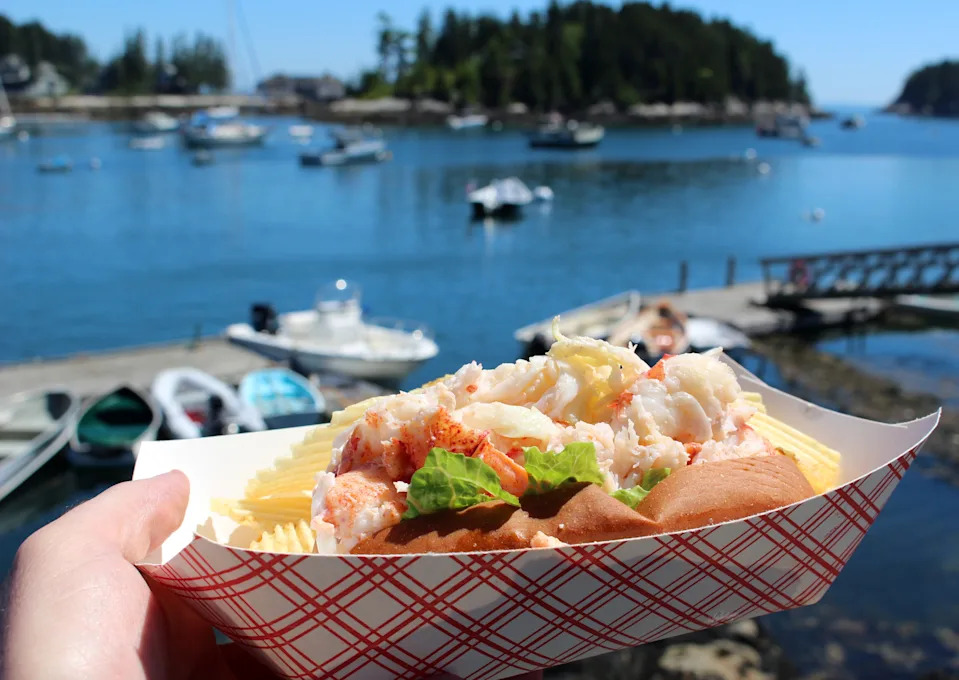 A lobster roll is held up against a scenic harbor backdrop with boats and calm water