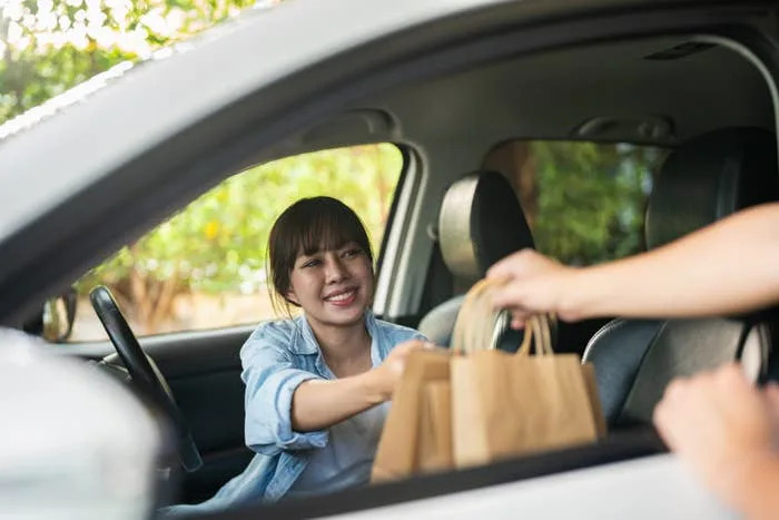 A person in a car receives a brown paper bag from another person outside, suggesting a food delivery or curbside pickup interaction