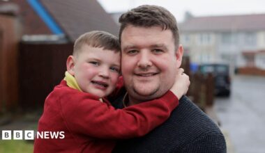A small boy puts his hands round a man with short brown hair. The boy has short brown hair and a red jumper and a yellow polo. The man has a black sweatshirt on. Behind them is an out-of-focus street. The sky is grey.