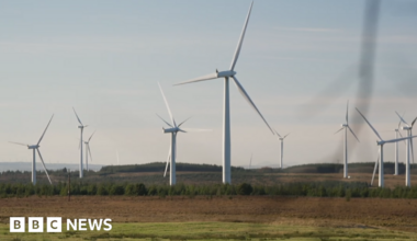 About 10 white wind turbines spinning on a green and brown field site with a grey sky overheard