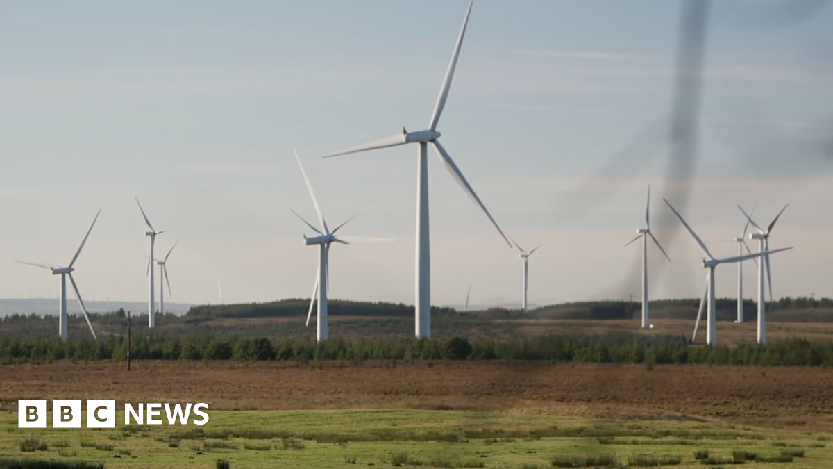 About 10 white wind turbines spinning on a green and brown field site with a grey sky overheard