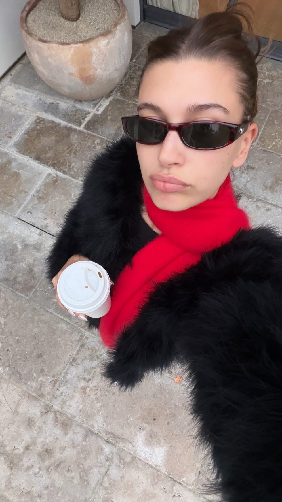 Hailey Bieber selfie person holding a coffee cup while wearing a black fur coat and red scarf