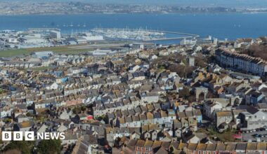 Aerial view across Portland looking towards Weymouth. In the foreground, taking up most of the photo, is the town of Fortuneswell, consisting of mostly terraced houses built on a hill. Beyond the houses is Portland Port and Portland Habour and a large marina full of small boats. On the other side of the harbour is the Wyke Regis area of Weymouth.
