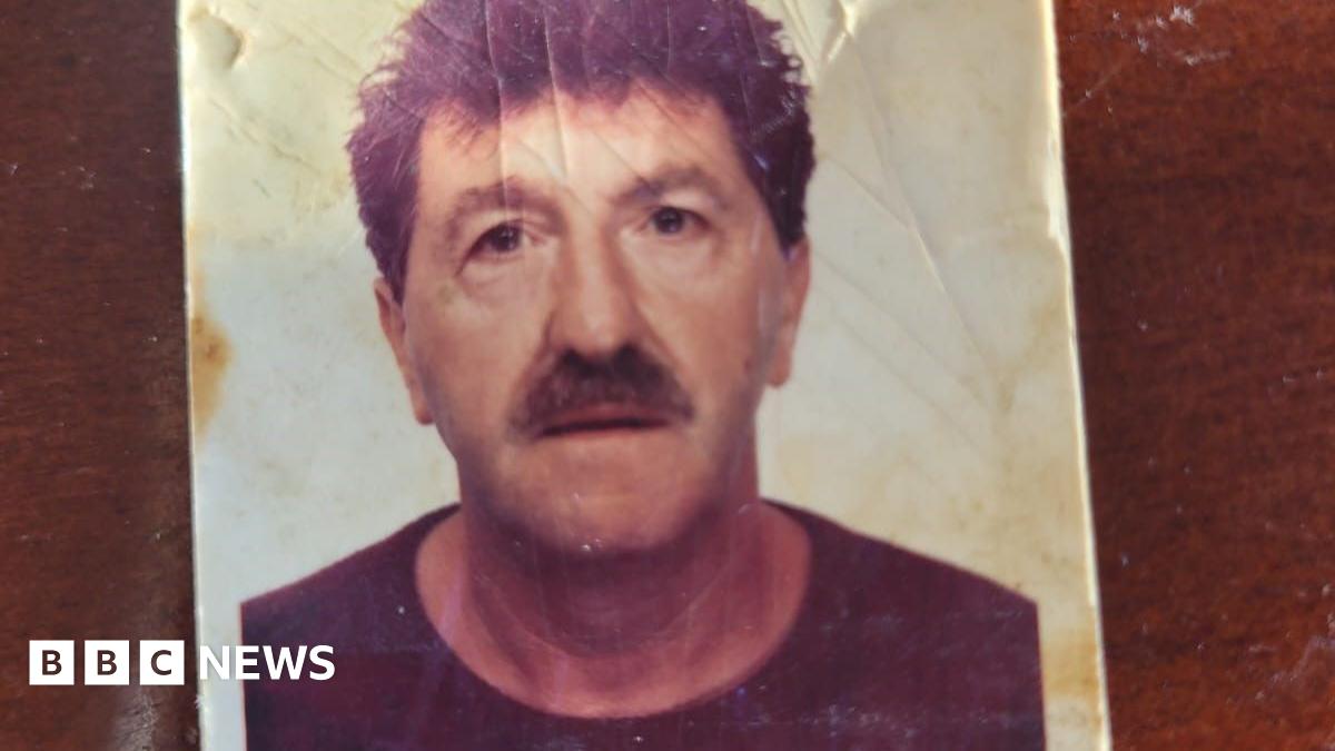 A passport-style creased photograph of a man with dark hair and a moustache. He is staring into the camera.