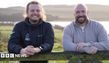 Jonny Snape, nature recovery ranger, and Matthew Squires, from The Squires Foundation. They are outdoors in an area of open countryside, with a lake in the background. They are both leaning on a gate or fence and smiling at the camera. Mr Snape is wearing a dark outdoor jacket, he has long blond curly hair tied back and a short beard. Mr Squires is bald with a trimmed brown beard. He is wearing a grey jumper.