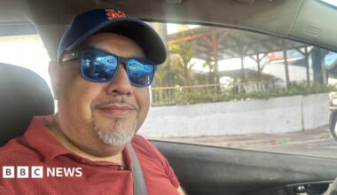 A man in the drivers seat of a car, wearing a blue cap and blue sun glasses looking into the camera.