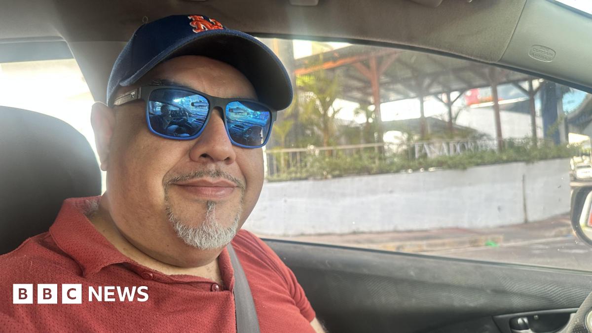 A man in the drivers seat of a car, wearing a blue cap and blue sun glasses looking into the camera.