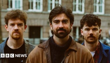 Oisín McCaffrey (a ginger haired man with a black leather jacket and black sweater), Robbie Cunningham (medium length brown hair and beard, with a black sweater and brown jacket) and Ross McNerney (also medium length brown hair, a moustache and blue jacket) - all pictured from the chest up with a brown brick building visible behind them