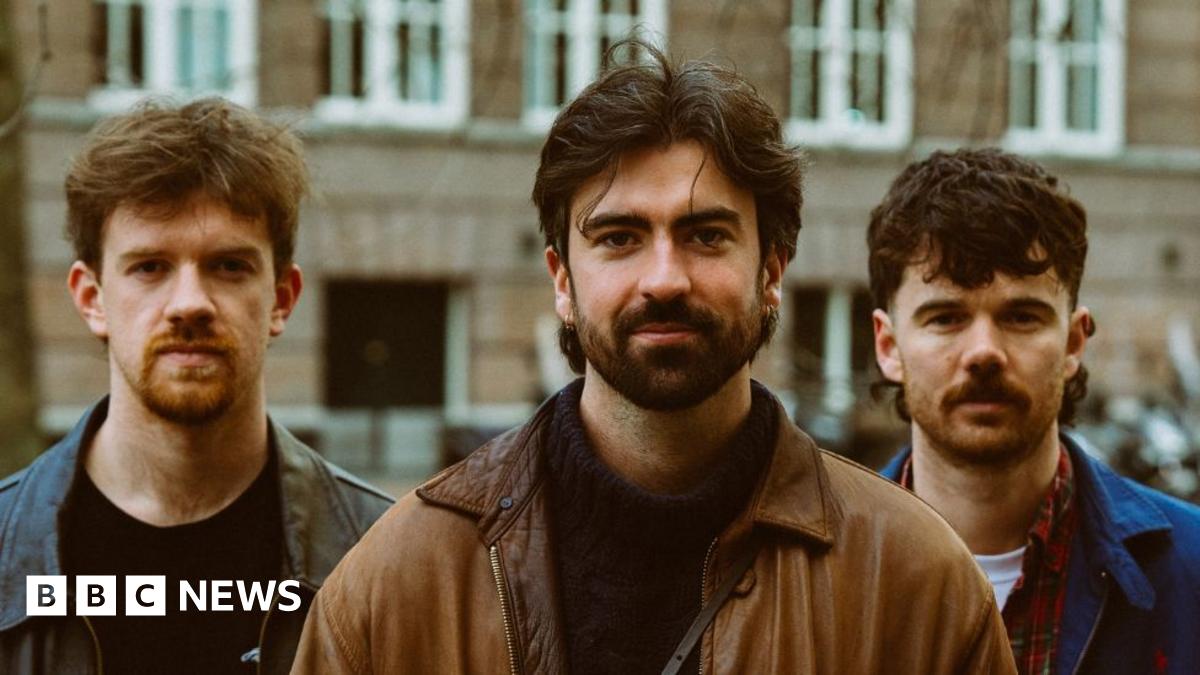 Oisín McCaffrey (a ginger haired man with a black leather jacket and black sweater), Robbie Cunningham (medium length brown hair and beard, with a black sweater and brown jacket) and Ross McNerney (also medium length brown hair, a moustache and blue jacket) - all pictured from the chest up with a brown brick building visible behind them