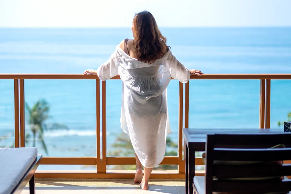 Person in relaxed pose on a balcony, overlooking the ocean. They wear a light, casual robe, suggesting leisure or a break from work