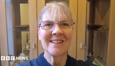 A woman has grey hair pulled back with a fringe and glasses. She is wearing a navy blue jumper with white spots and standing smiling in front of a cabinet