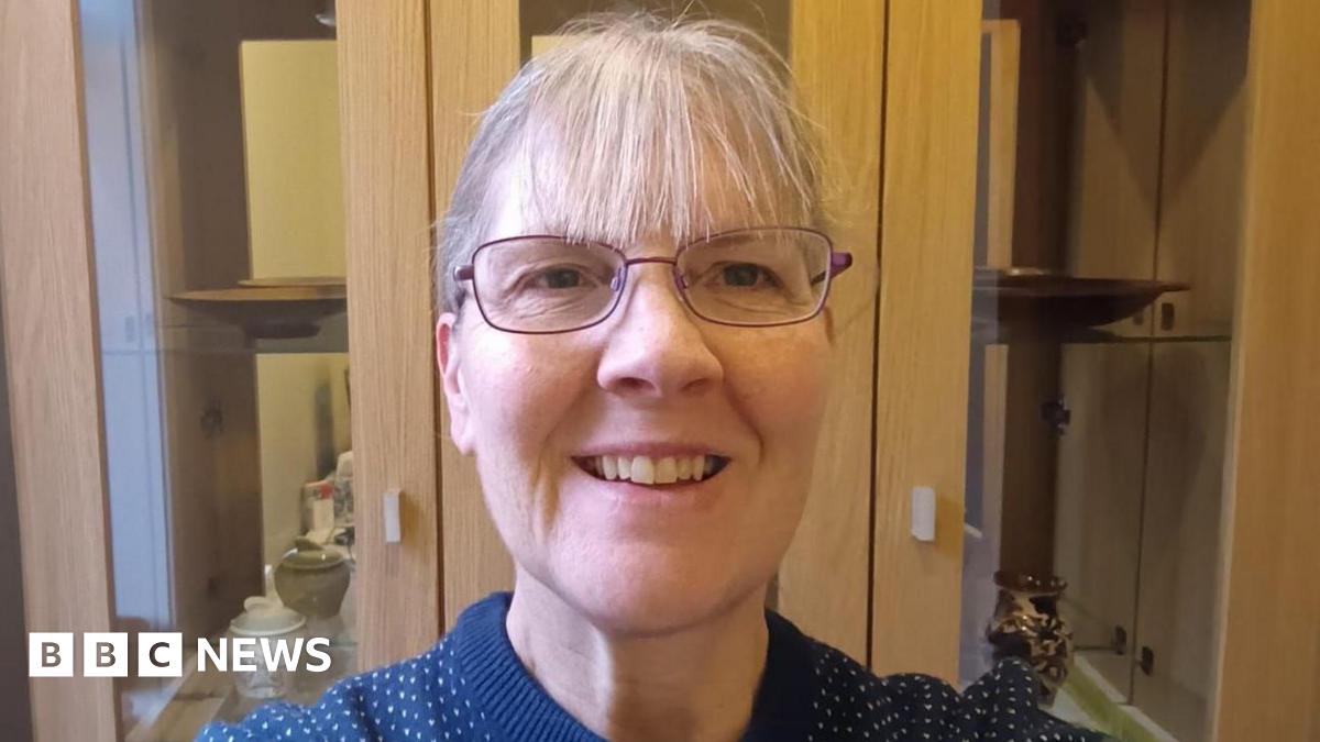 A woman has grey hair pulled back with a fringe and glasses. She is wearing a navy blue jumper with white spots and standing smiling in front of a cabinet