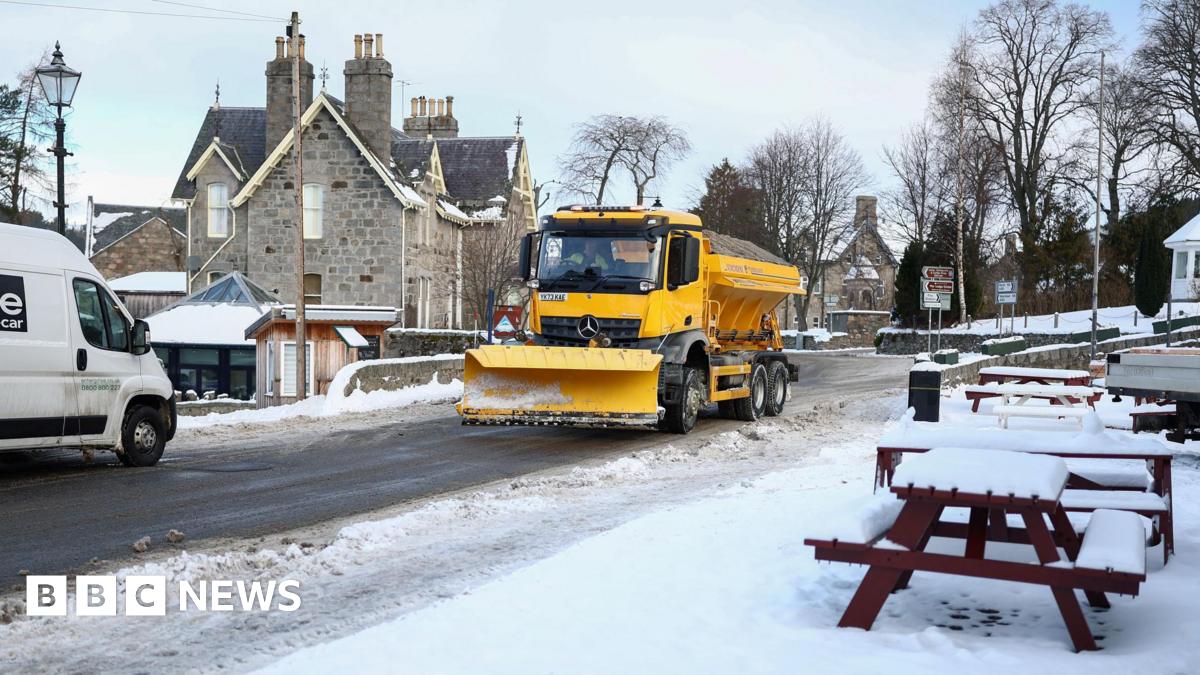 A yellow gritting lorry fitted with a snow plough travels down a snowy street in the picturesque village of Braemar.