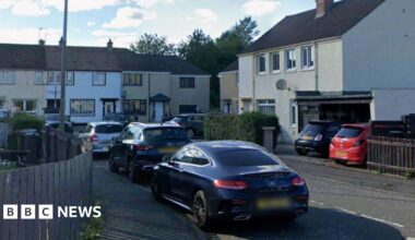 A cul-de-sac with brown and white houses. There are trees behind the houses at the far end. There are three cars in the foreground.
