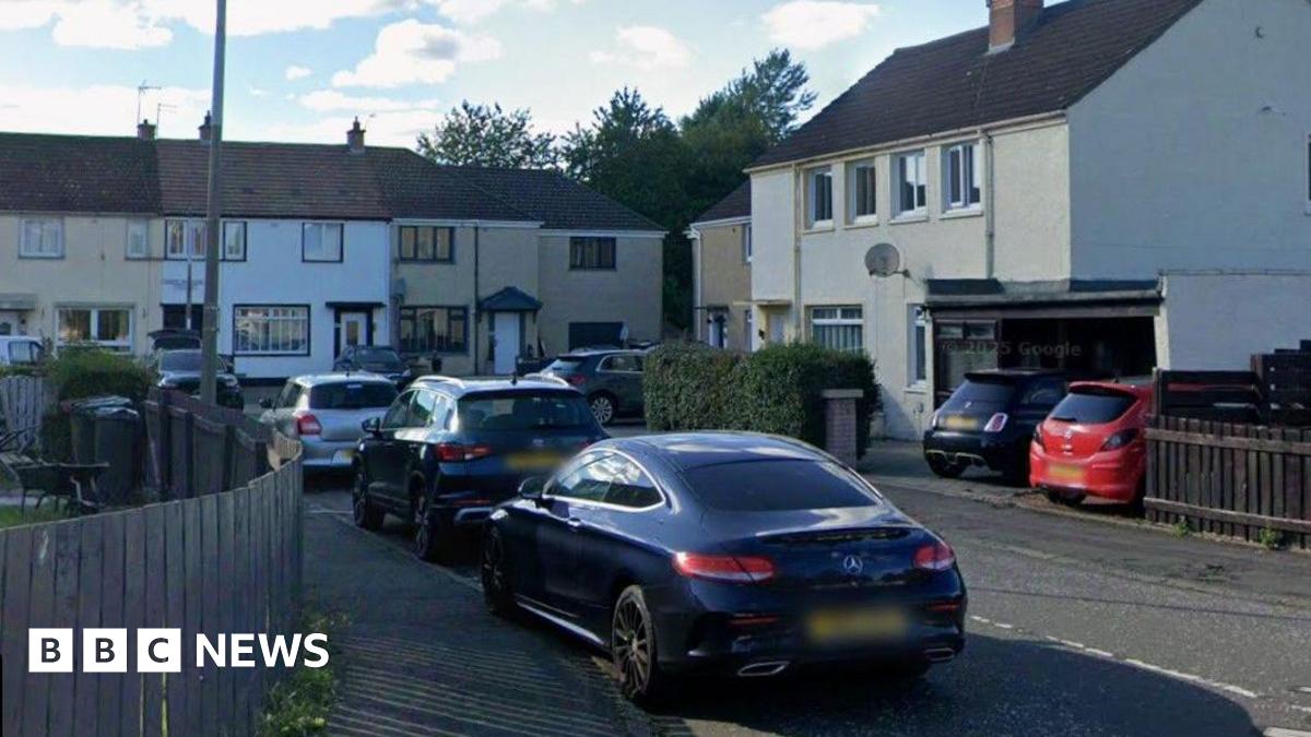A cul-de-sac with brown and white houses. There are trees behind the houses at the far end. There are three cars in the foreground.