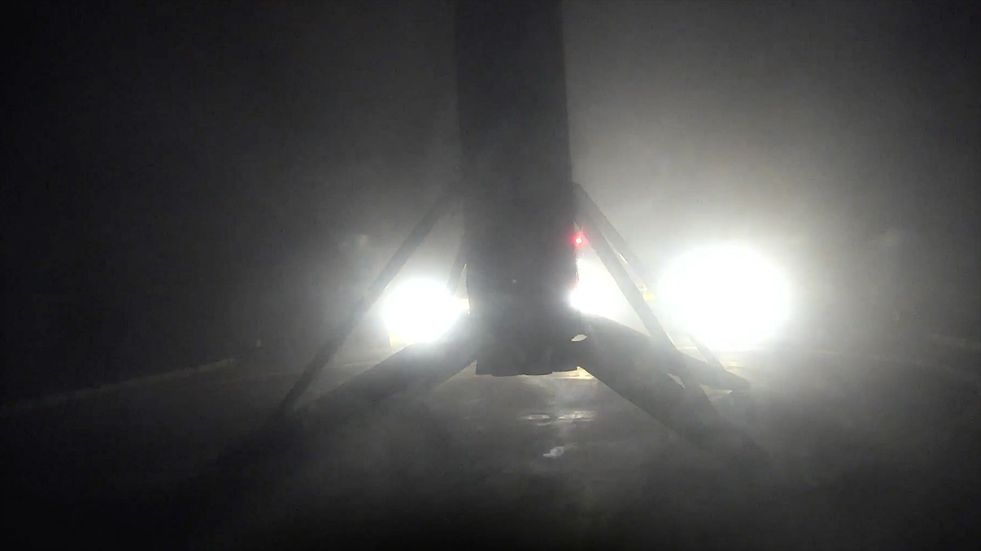 the first stage of an orbital rocket is seen in silhouette, backlit by spotlights on the deck of a ocean-based landing platform