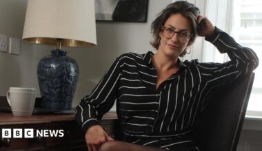Madelaine Thomas is sitting at a desk, turned towards the camera smiling. She is wearing a black and white striped shirt dress and sheer black tights. She has short dark hair with a blonde streak at the front and is wearing oversized reading glasses. On the desk there is a lamp with a blue base and a cream shade and a white mug.