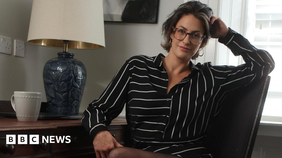 Madelaine Thomas is sitting at a desk, turned towards the camera smiling. She is wearing a black and white striped shirt dress and sheer black tights. She has short dark hair with a blonde streak at the front and is wearing oversized reading glasses. On the desk there is a lamp with a blue base and a cream shade and a white mug.
