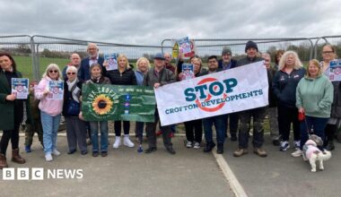 A group of people including men and women stood in a line. They are holding posters, one which reads 'Stop Crofton Development'. In the background is a green field and metal barriers.