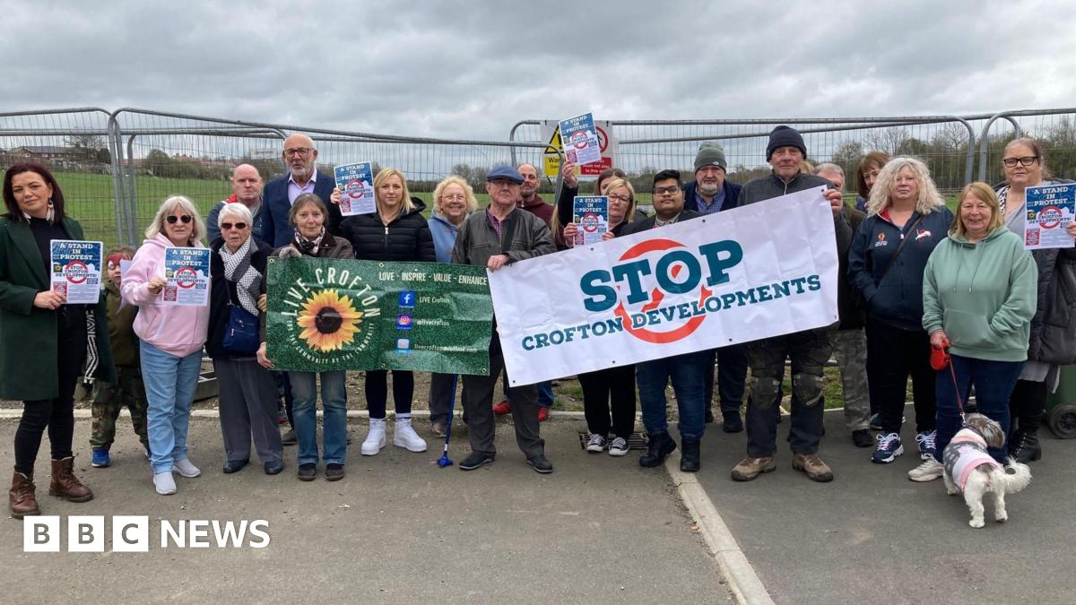 A group of people including men and women stood in a line. They are holding posters, one which reads 'Stop Crofton Development'. In the background is a green field and metal barriers.