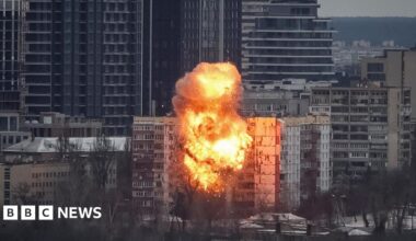 Orange fire and smoke rise from a multi-storey building after it is hit by a missile in Kyiv on 27 December. Taller buildings are situated behind the one targeted, with trees in front of it.