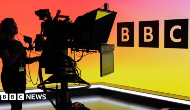 A woman operating a TV camera in front of the BBC Logo on an LED wall.