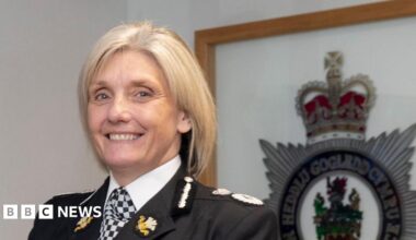 Amanda Blakeman in police uniform smiling at the camera. She has blonde bobbed hair and stands in front of framed North Wales Police crest