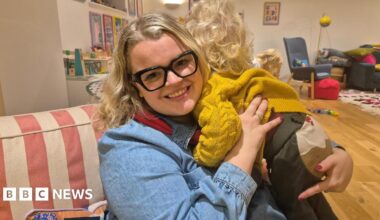 A blonde woman with black, square glasses holding a blonde male toddler over her shoulder