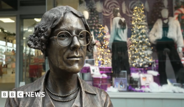 A close up of the head and shoulders of the bronze statue with the shop windows in the background. The subject has curly ear-length hair, round glasses and two strings of pearls or beads around her neck.