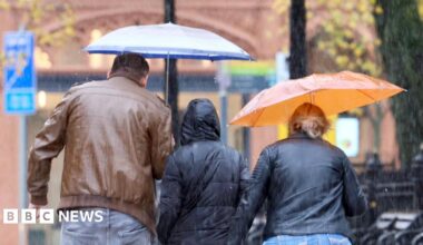 A man and woman are walking on a pavement in Belfast City Centre. It is raining and she is holding a clear umbrella over her head. The man has his hood up. A light blue car on the street is driving through a very large puddle and the water on the road is splashing up and hitting the couple on the pavement.