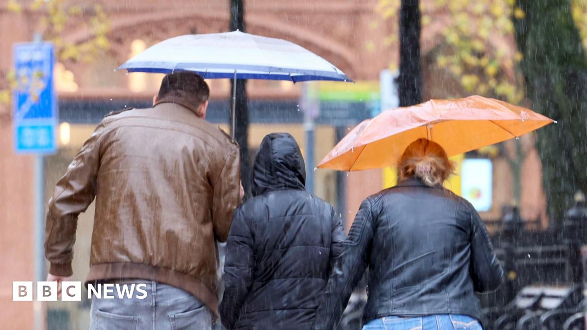 A man and woman are walking on a pavement in Belfast City Centre. It is raining and she is holding a clear umbrella over her head. The man has his hood up. A light blue car on the street is driving through a very large puddle and the water on the road is splashing up and hitting the couple on the pavement.