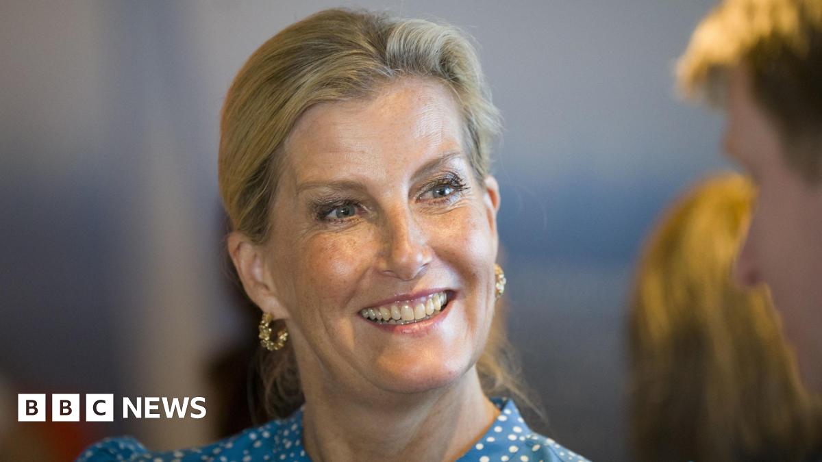 The Duchess of Edinburgh is smiling at someone off camera. She has blonde hair tied back from her face and blue eyes. She is wearing ornate gold hoop earrings and a blue and white spotted top.