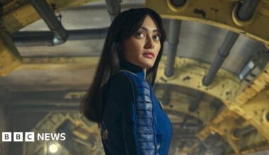 A woman with long black hair stands in the middle of a steampunk-like room. There are pipes and girders visible with yellow and grey colours. The woman has wide eyes and is wearing a deep blue jumpsuit.