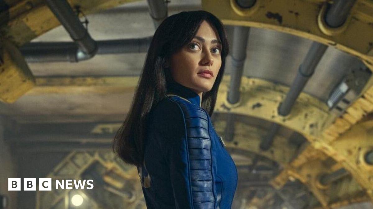 A woman with long black hair stands in the middle of a steampunk-like room. There are pipes and girders visible with yellow and grey colours. The woman has wide eyes and is wearing a deep blue jumpsuit.