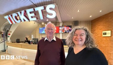 John Sambrook, a man wearing glasses and a maroon coloured jumper. He is standing next to a brick-building with white lettering on the side which reads New Vic Theatre.