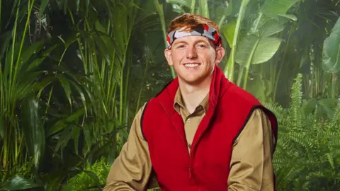 ITV AngryGinge wears a red vest and tan shirt and a headband in front of a wall with forest leaves on it.