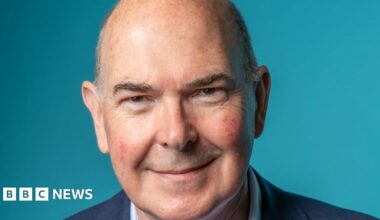 A bald, older man wearing a navy blue suit smiles against a lighter blue background.
