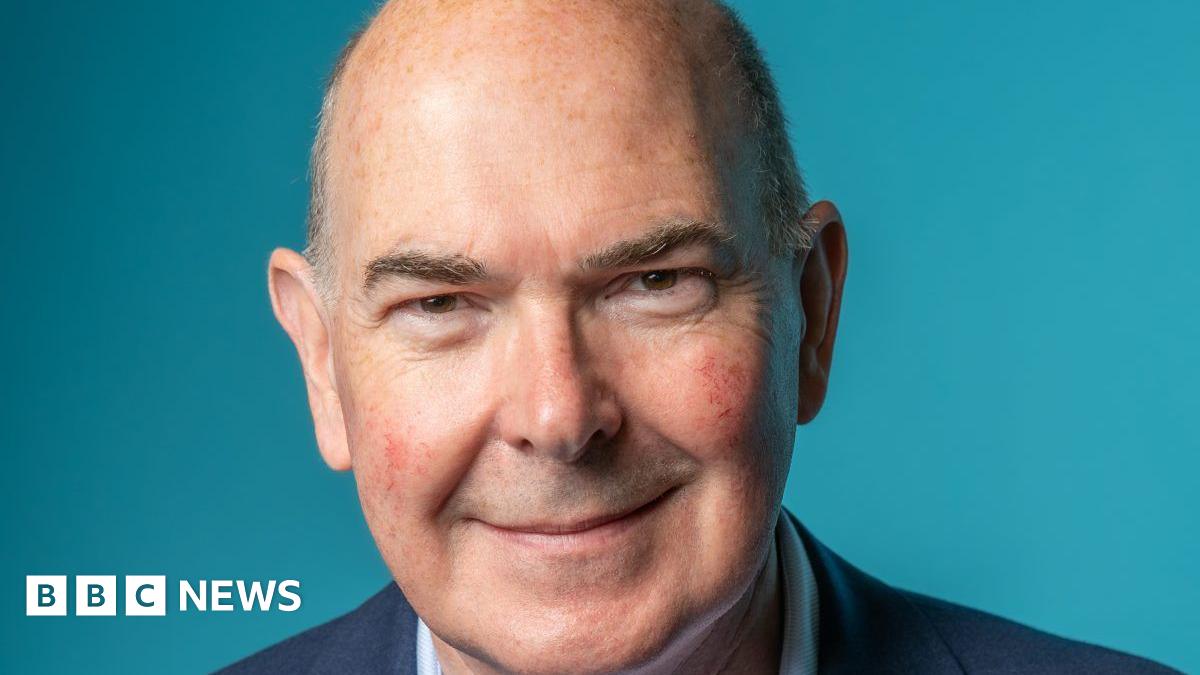 A bald, older man wearing a navy blue suit smiles against a lighter blue background.