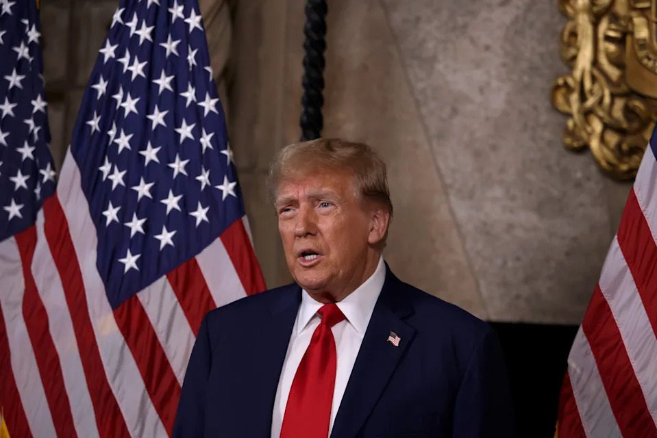 I don’t know who this is, but a person in a suit and red tie speaks at an event, standing in front of several U.S. flags