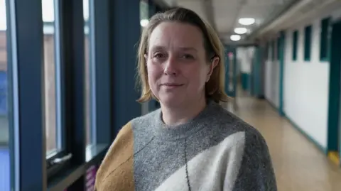 Gemma Laister/BBC A lady stands in a corridor smiling at the camera, She is wearing a grey and brown jumper. 