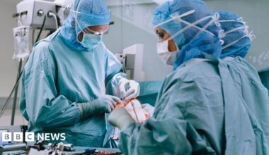 A group of doctors wearing blue scrubs, masks and hairnets. They are in a surgical theatre performing a knee replacement operation on a patient.